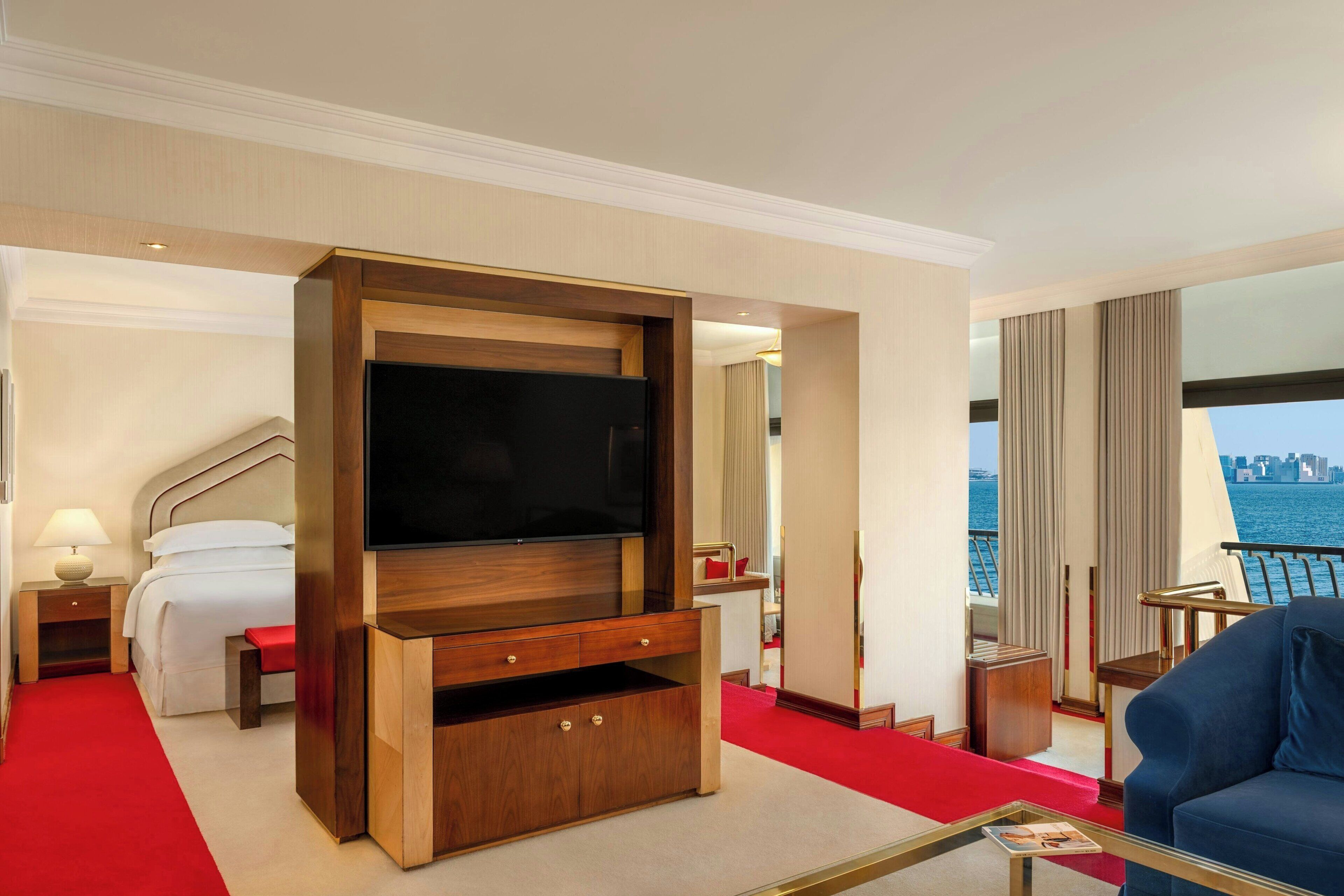 Quarto de hotel com televisão grande, cama, tapete vermelho e vista para a água.