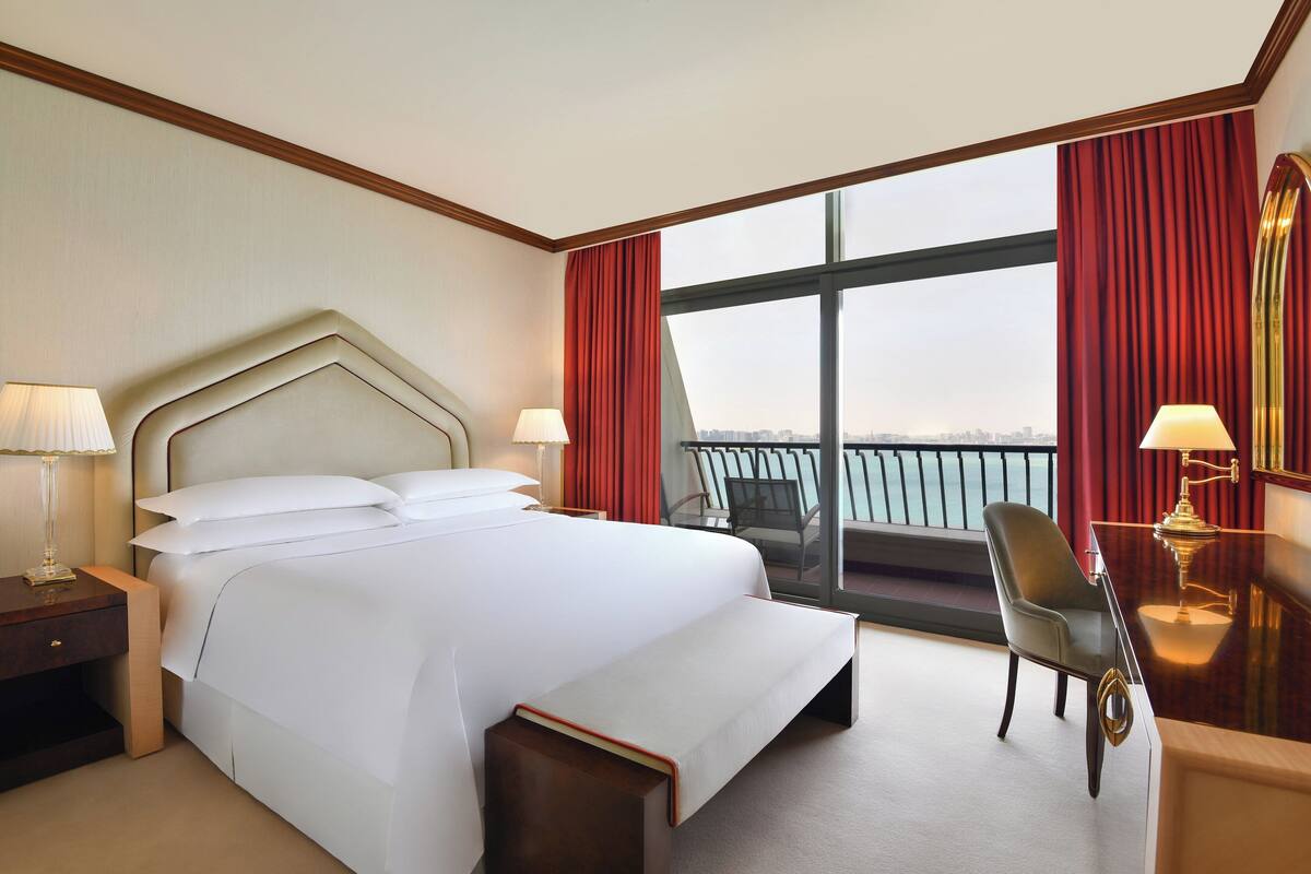A hotel room with a large bed, a desk with a chair, and a view of the city through large windows.
