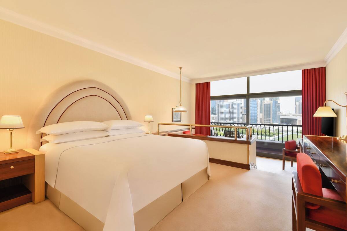A hotel room with a large bed, a desk, a chair, a TV, and a balcony with a city view.