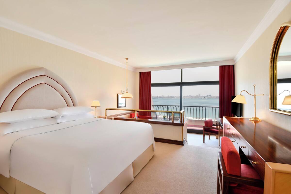 A hotel room with a large bed, a desk, a chair, a desk lamp, a bedside table, a mirror, and a view of the city through a window.