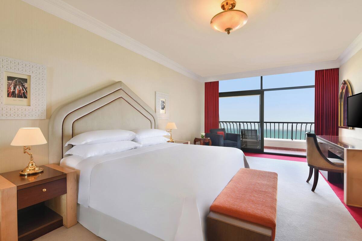 A hotel room with a large bed, a desk with a lamp, a chair, a TV, and a balcony with a view.