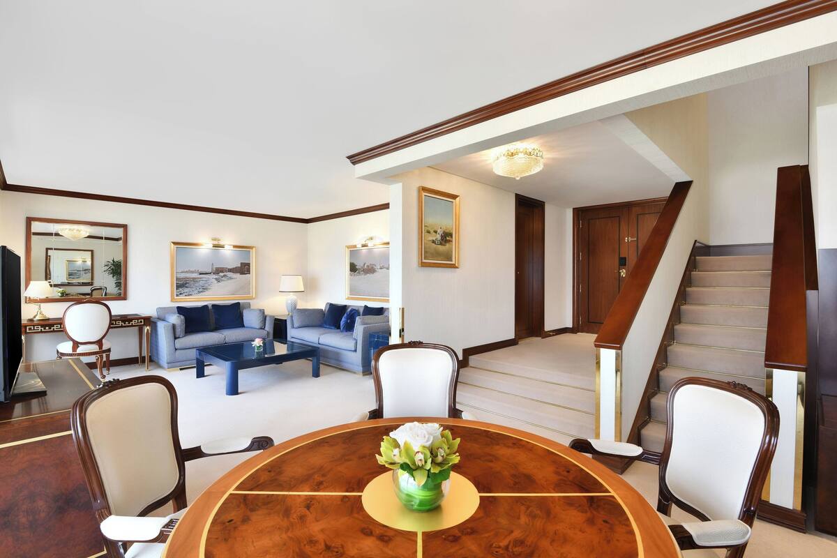 A spacious living room with a round wooden table, comfortable seating, a staircase, and a large painting on the wall.