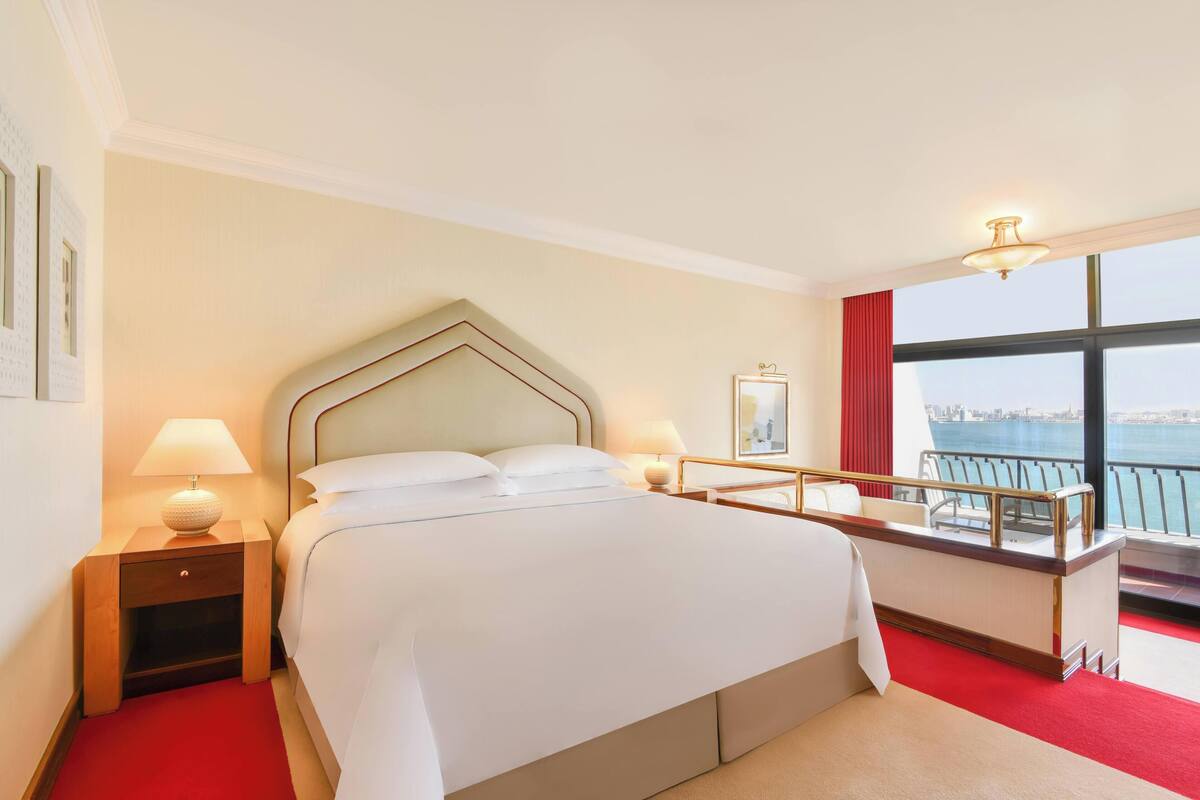 A hotel room with a large bed, bedside lamps, a nightstand, a desk with a chair, and a view of the water.