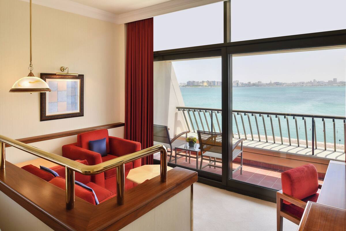A hotel room with a balcony overlooking a body of water, a red sofa, a wooden desk, and a framed picture on the wall.