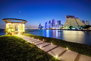 Vista al agua con un camino que lleva a un edificio de estructura de techo singular, rodeado de agua y un horizonte urbano iluminado.
