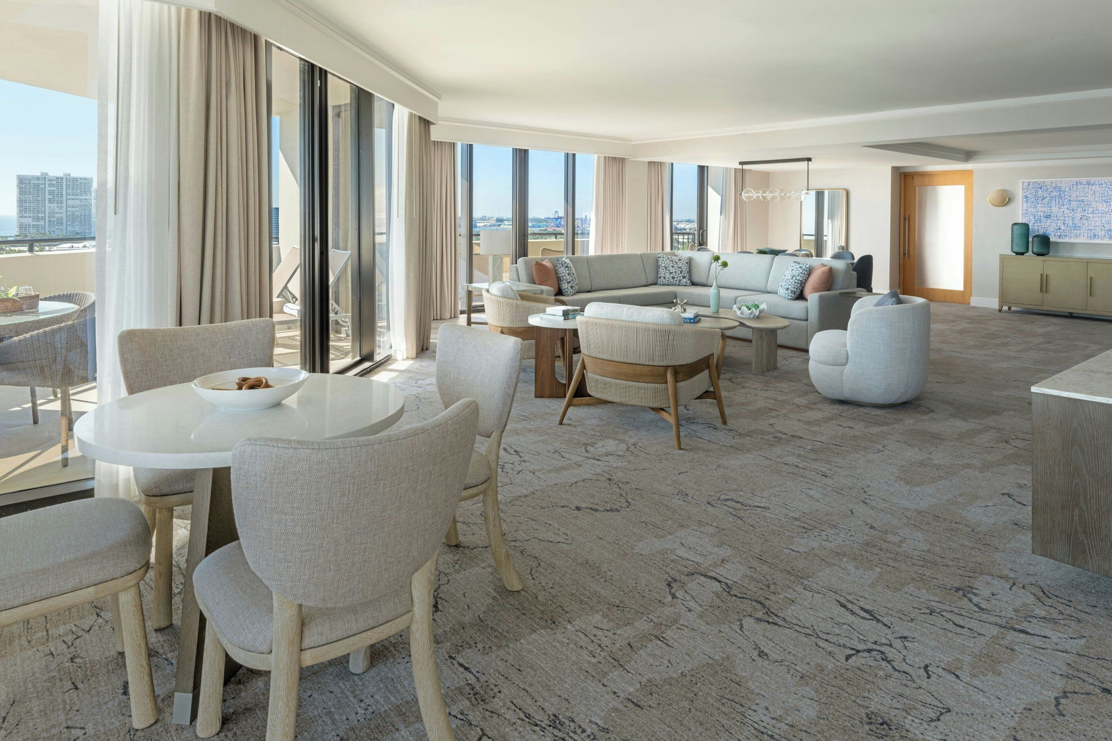 A modern hotel room with a large balcony, a dining area with a round table and chairs, a sofa set, and a large window offering a city view.