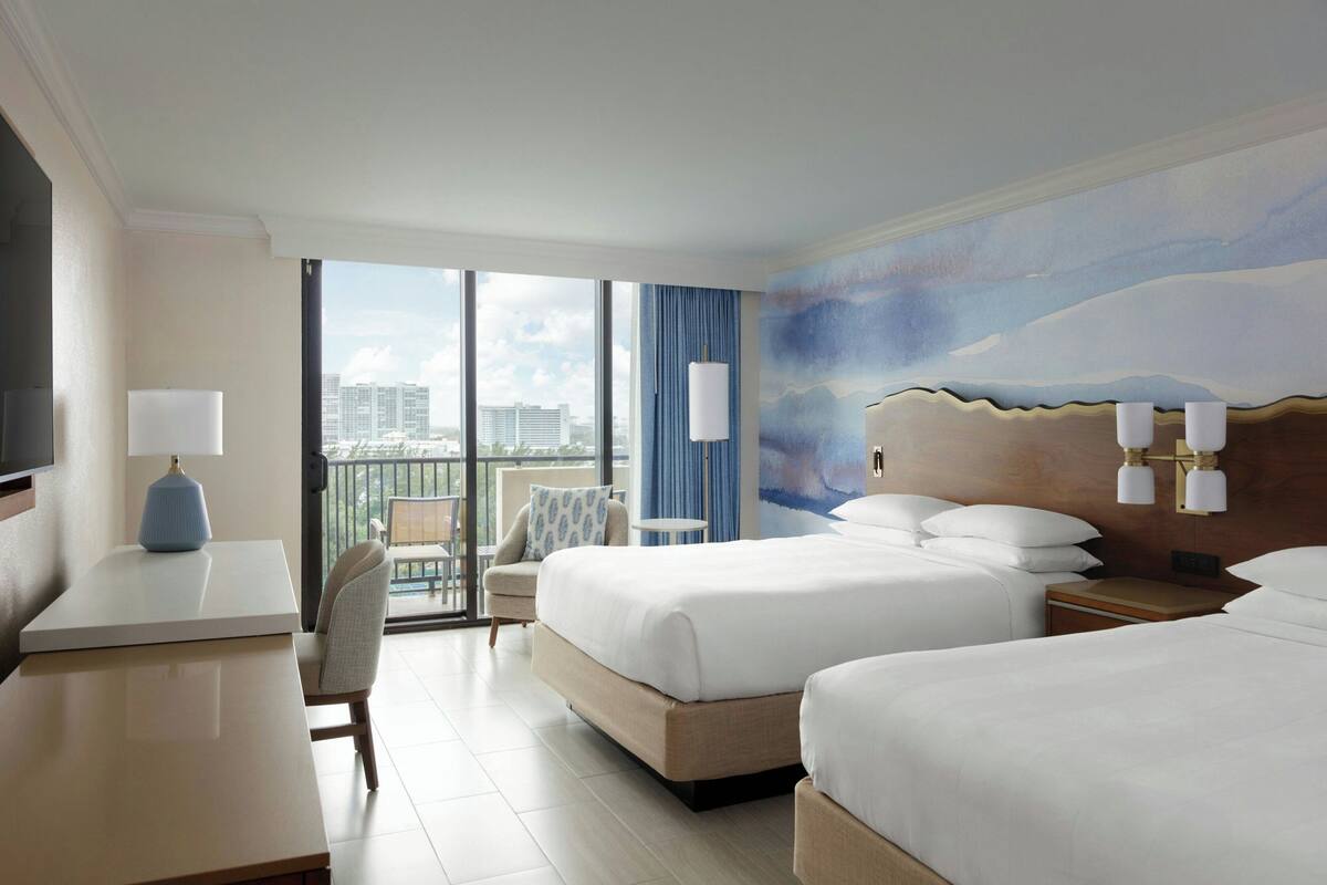Een hotelkamer met twee bedden, een bureau, een stoel, een televisie en een balkon met uitzicht op de stad.
