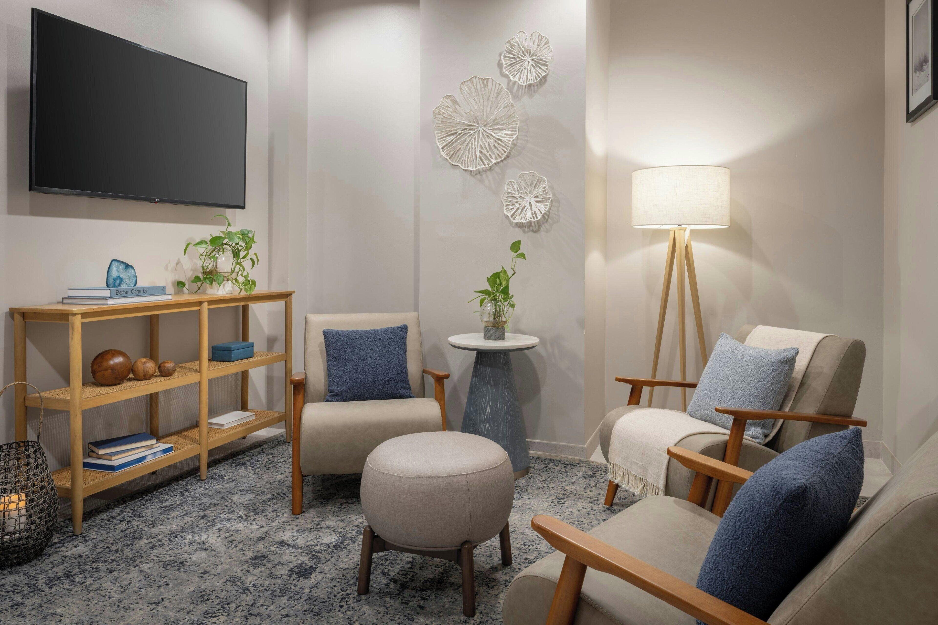 Una sala de estar con un televisor de pantalla plana, dos sillones, un otoman redondo, una mesita de madera y decoración en la pared.
