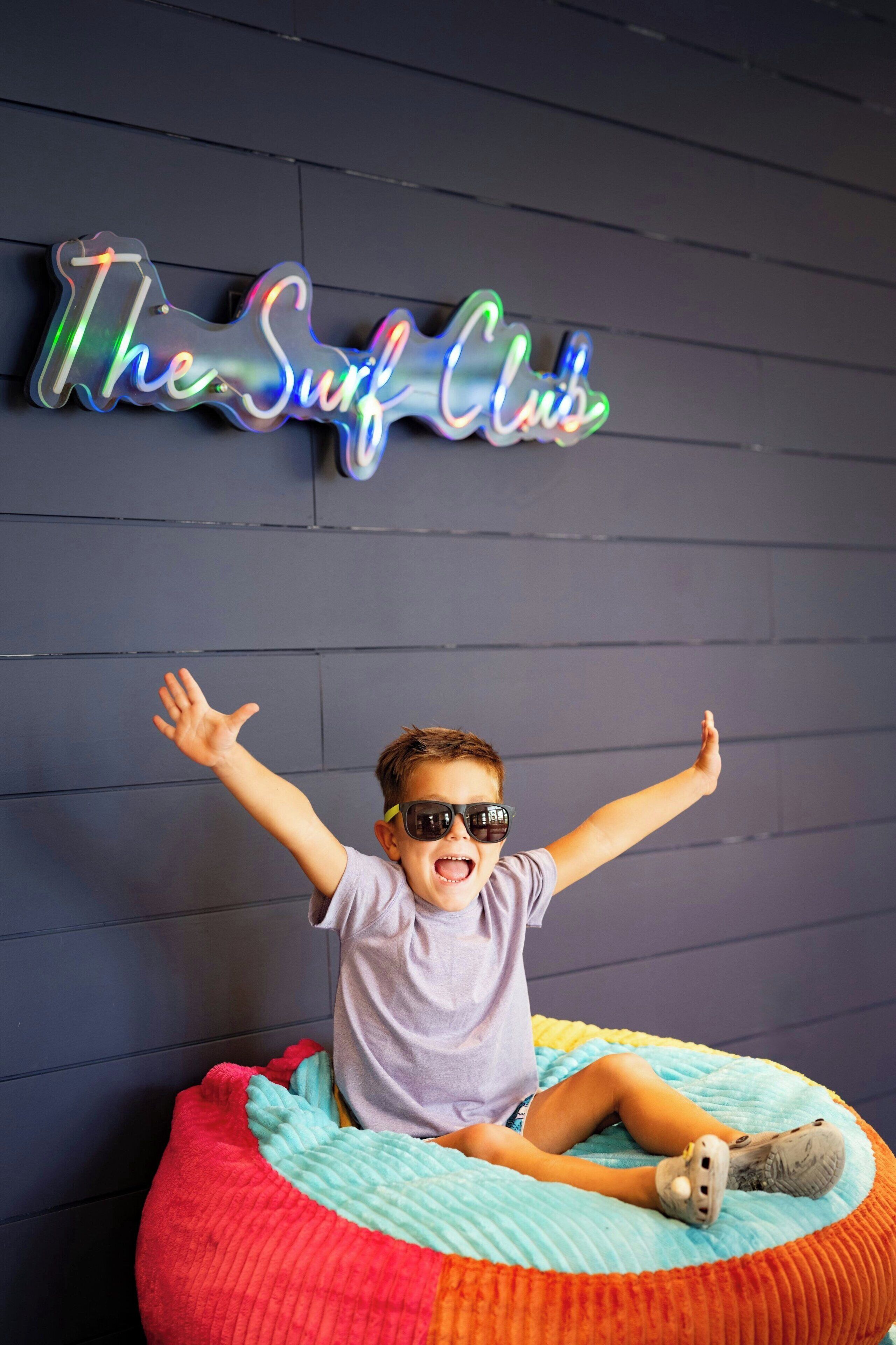 Un niño sentado en un sillón de peluche colorido con los brazos levantados, frente a un letrero de neón que dice 'The Surf Club'.