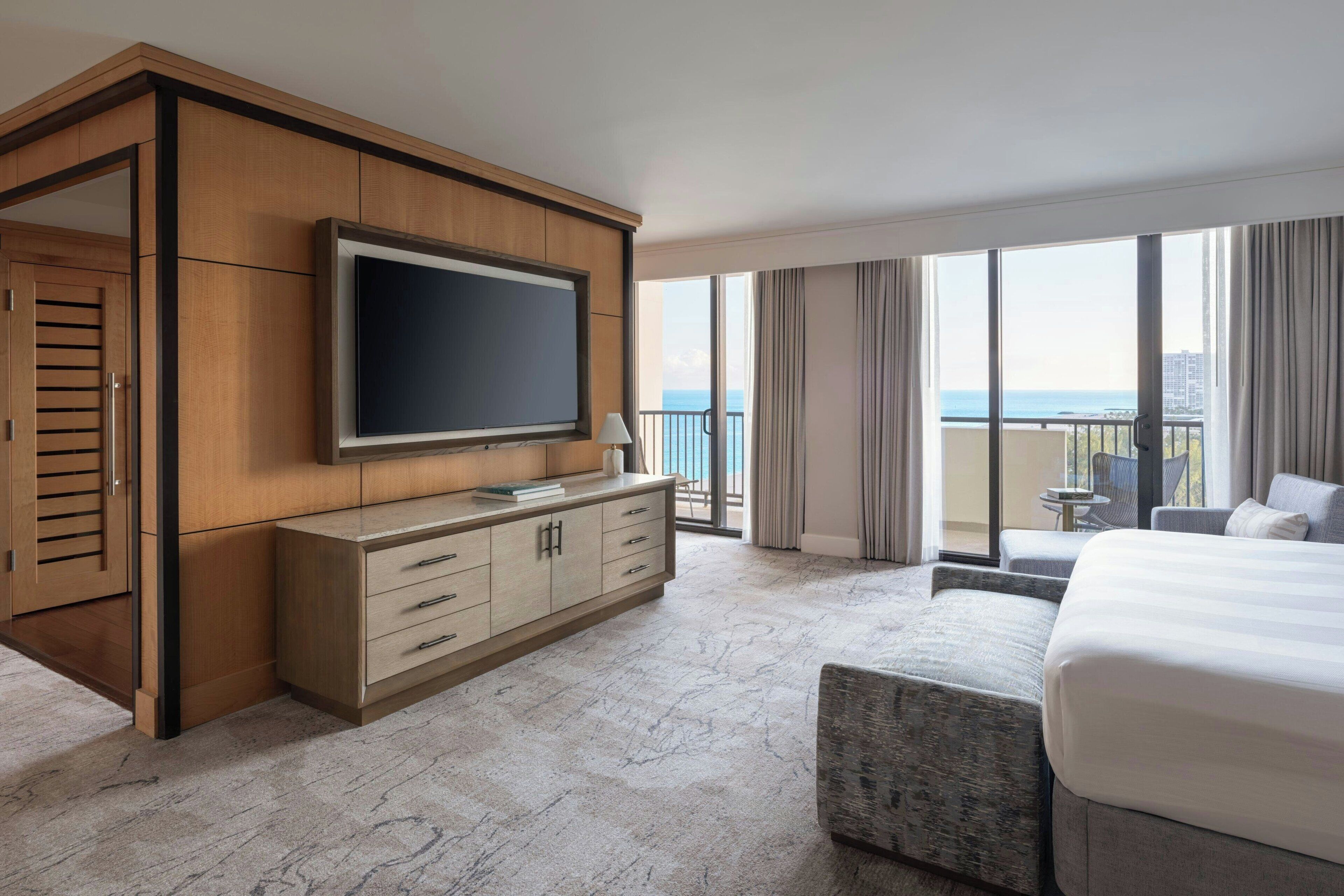 Une chambre d’hôtel avec un grand lit, un téléviseur posé sur une commode en bois, un balcon avec vue sur la mer et une petite table avec des chaises.