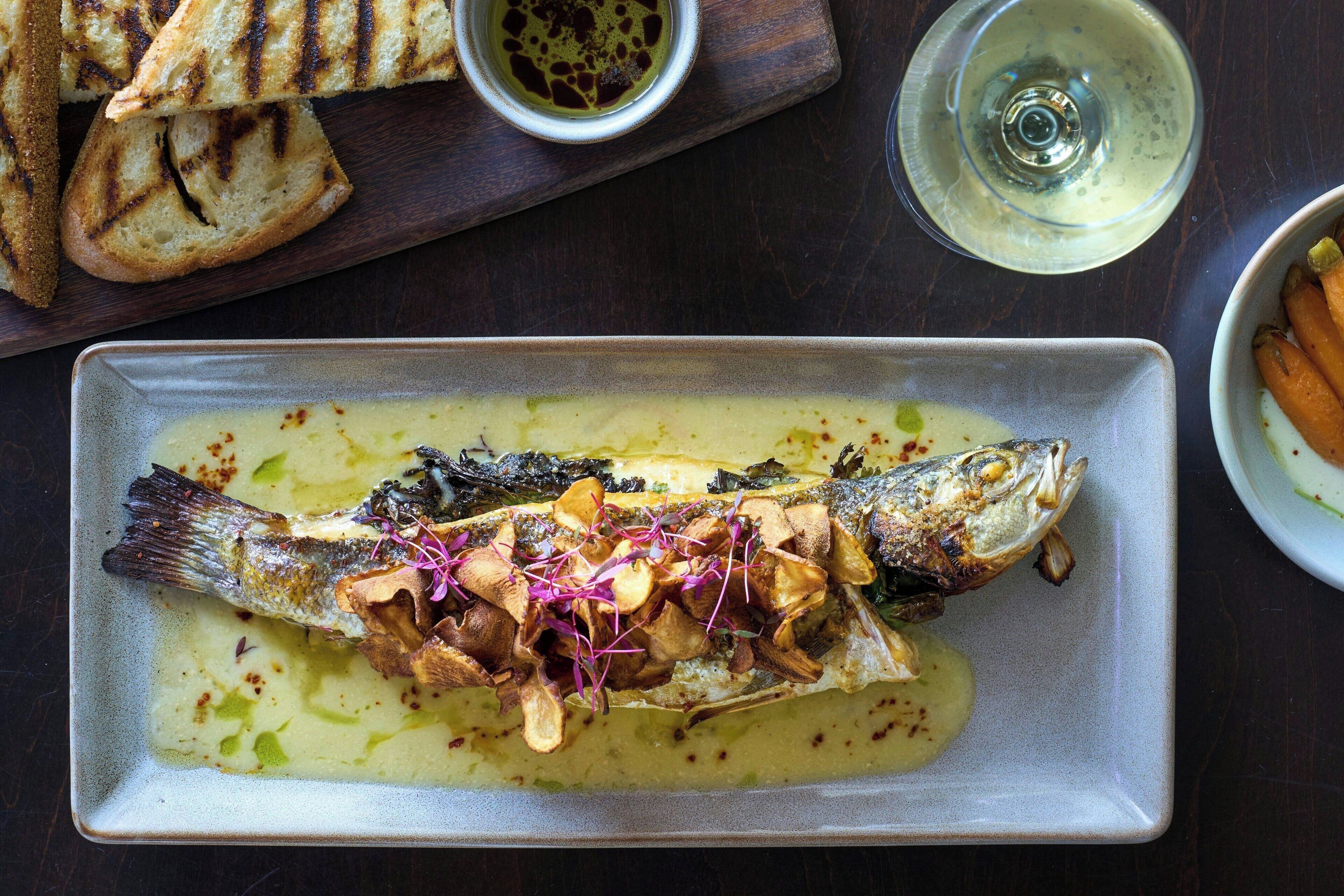 Plato con un guiso de pescado, setas y hierbas, acompañado de pan a la parrilla y una copa de vino blanco.