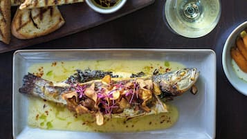 Plato con un guiso de pescado, setas y hierbas, acompañado de pan a la parrilla y una copa de vino blanco.