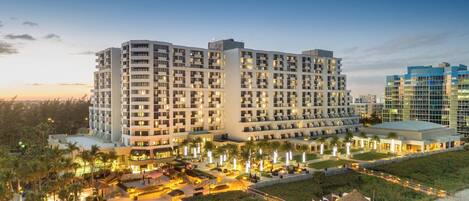 A multi-story hotel with a central courtyard, surrounded by palm trees and a beach view.