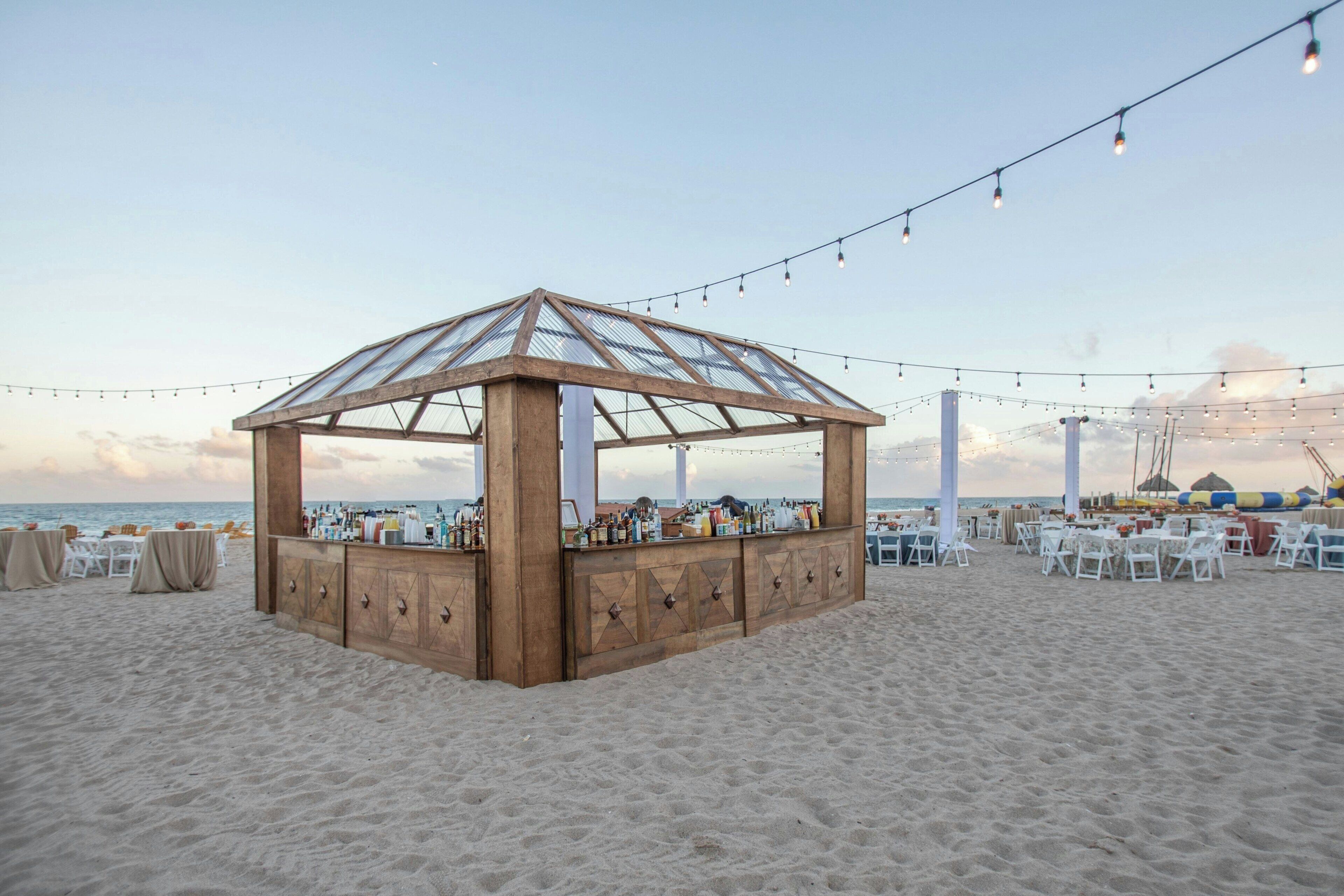A beachside gazebo with a bar, surrounded by tables and chairs, overlooking the sea.
