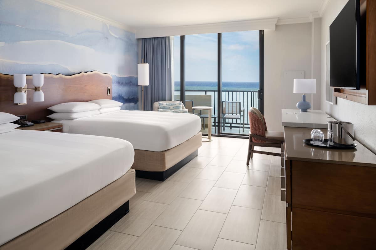 Een hotelkamer met twee bedden, een bureau, een stoel, een kleine tafel en een balkon met uitzicht op zee.