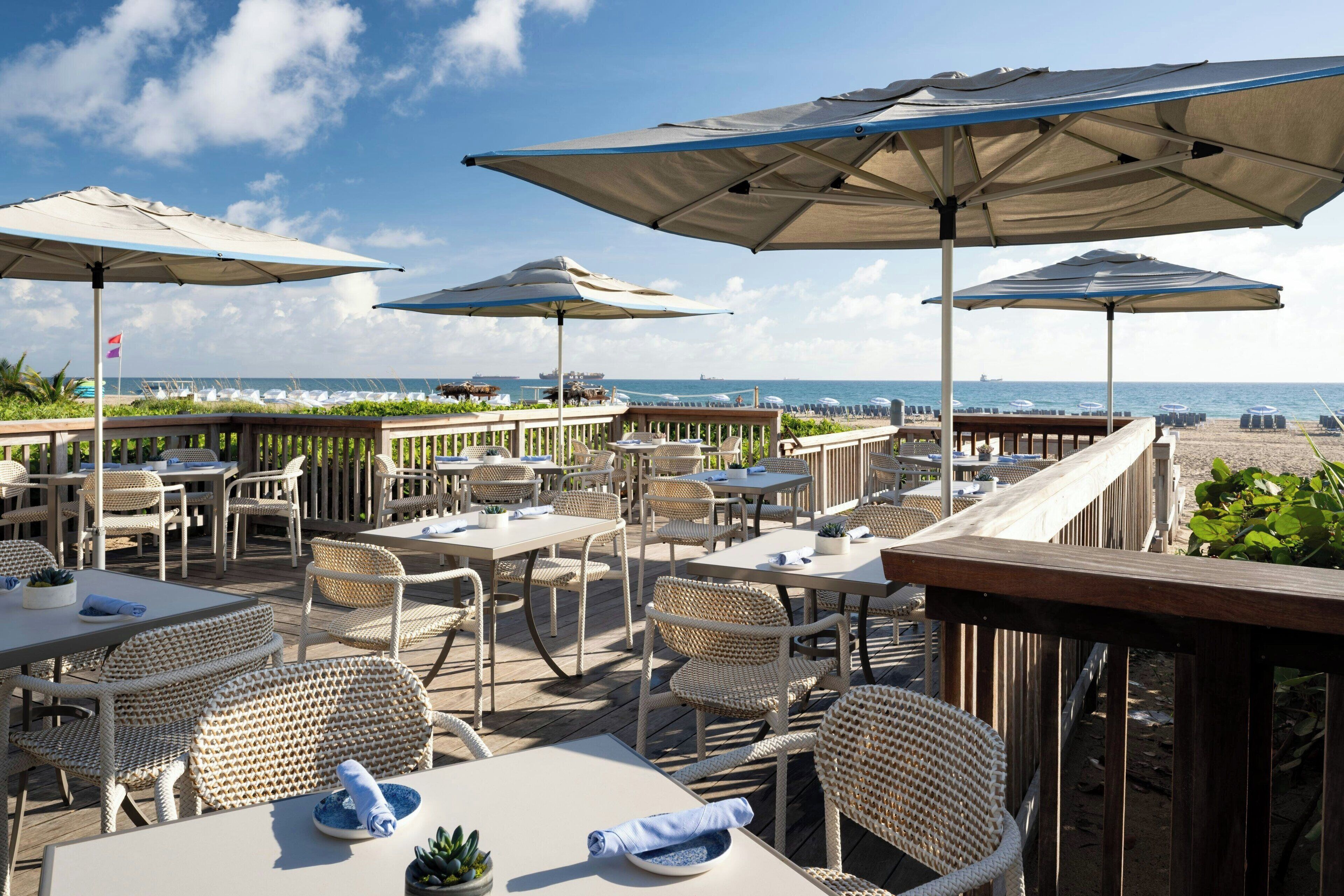 Un área de comedor al aire libre con mobiliario de mimbre y sombrillas, con vista a la playa.