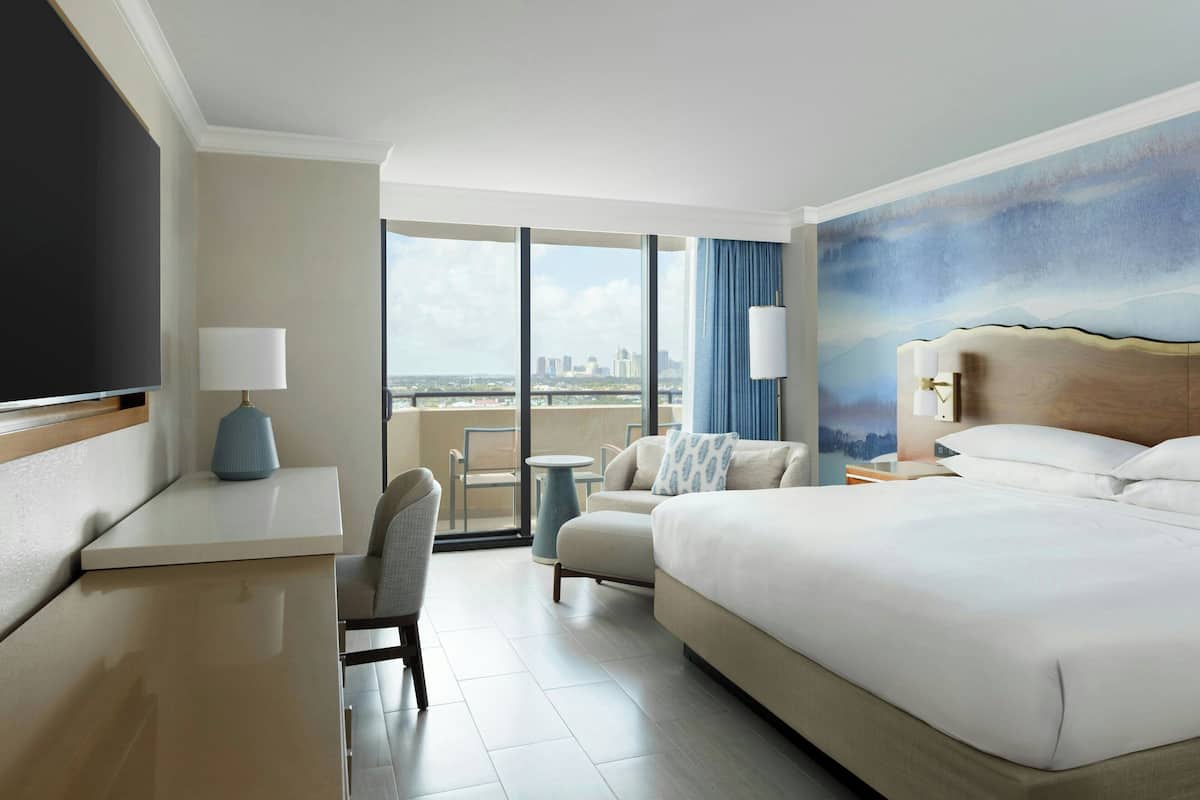Hotelkamer met een groot bed, een bureau, een stoel, een kleine tafel en uitzicht op de stad door grote ramen.