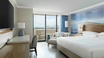 Habitación de hotel con una cama grande, un escritorio, una silla, una mesita y vistas a la ciudad a través de amplios ventanales.