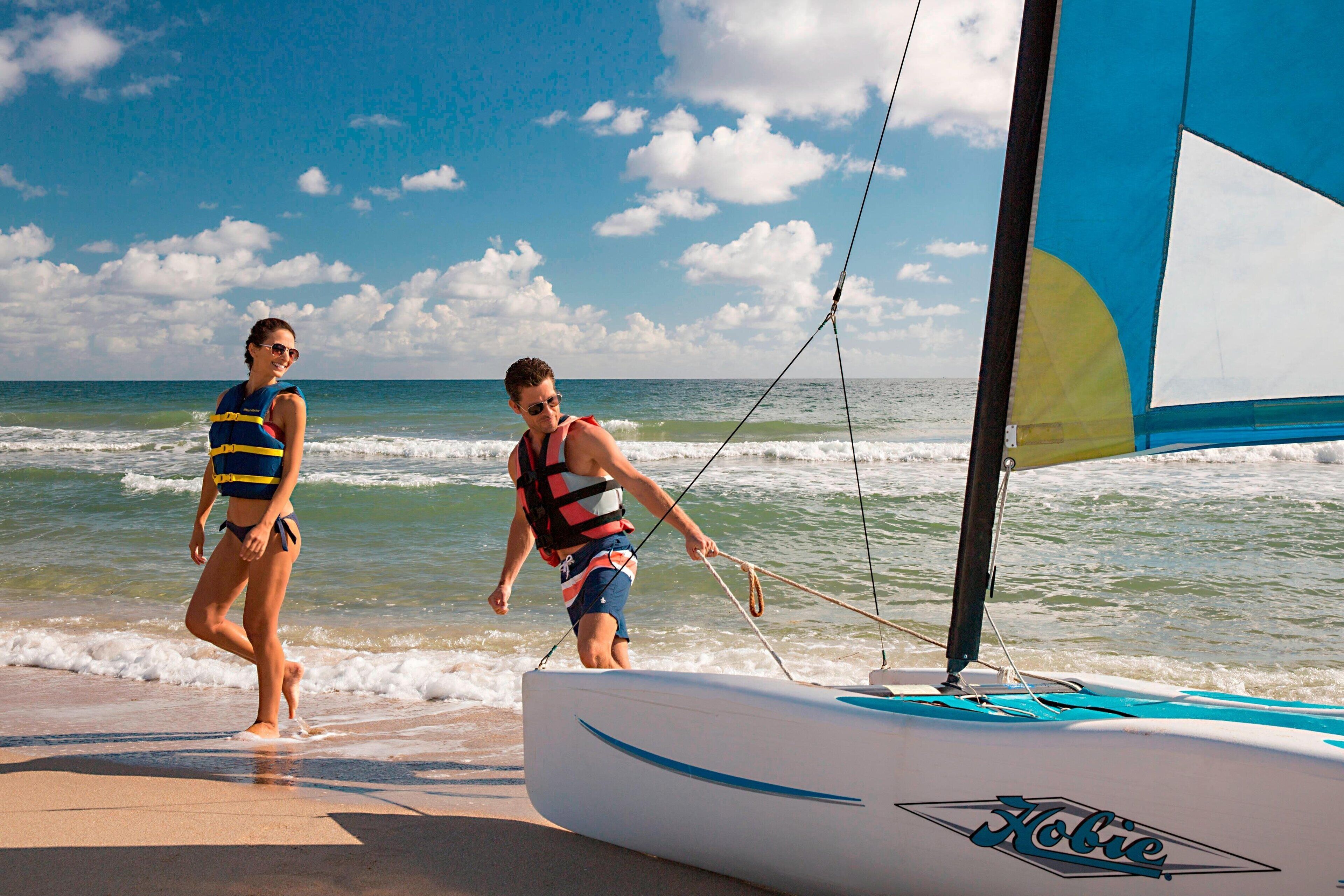 Un velero llamado 'Hobie' en una playa con dos personas usando chalecos salvavidas.