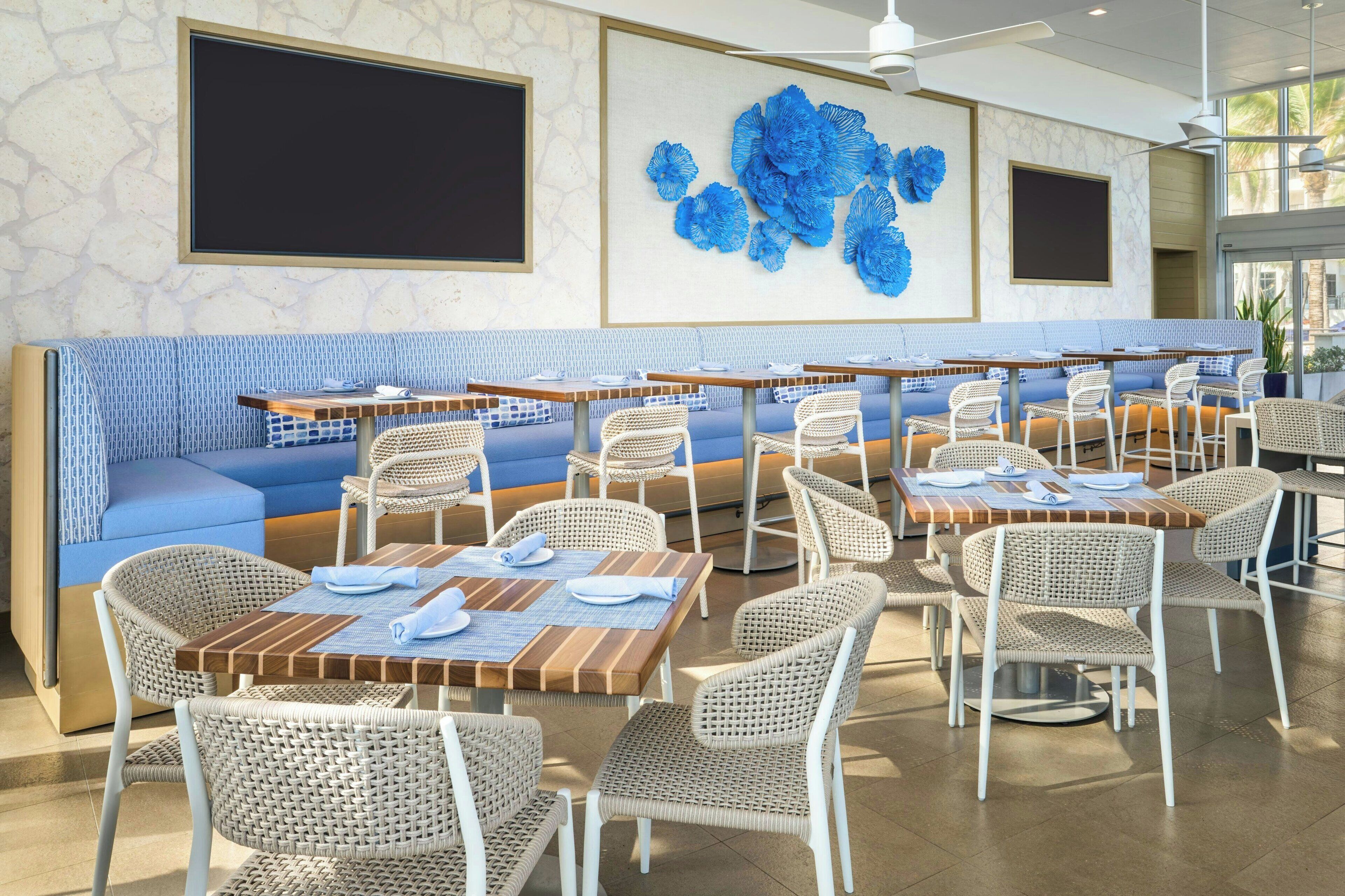 A restaurant with a long blue bench seating area, multiple tables with wicker chairs, and a large blue floral wall art piece.