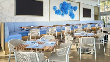 A restaurant with a long blue bench seating area, multiple tables with wicker chairs, and a large blue floral wall art piece.