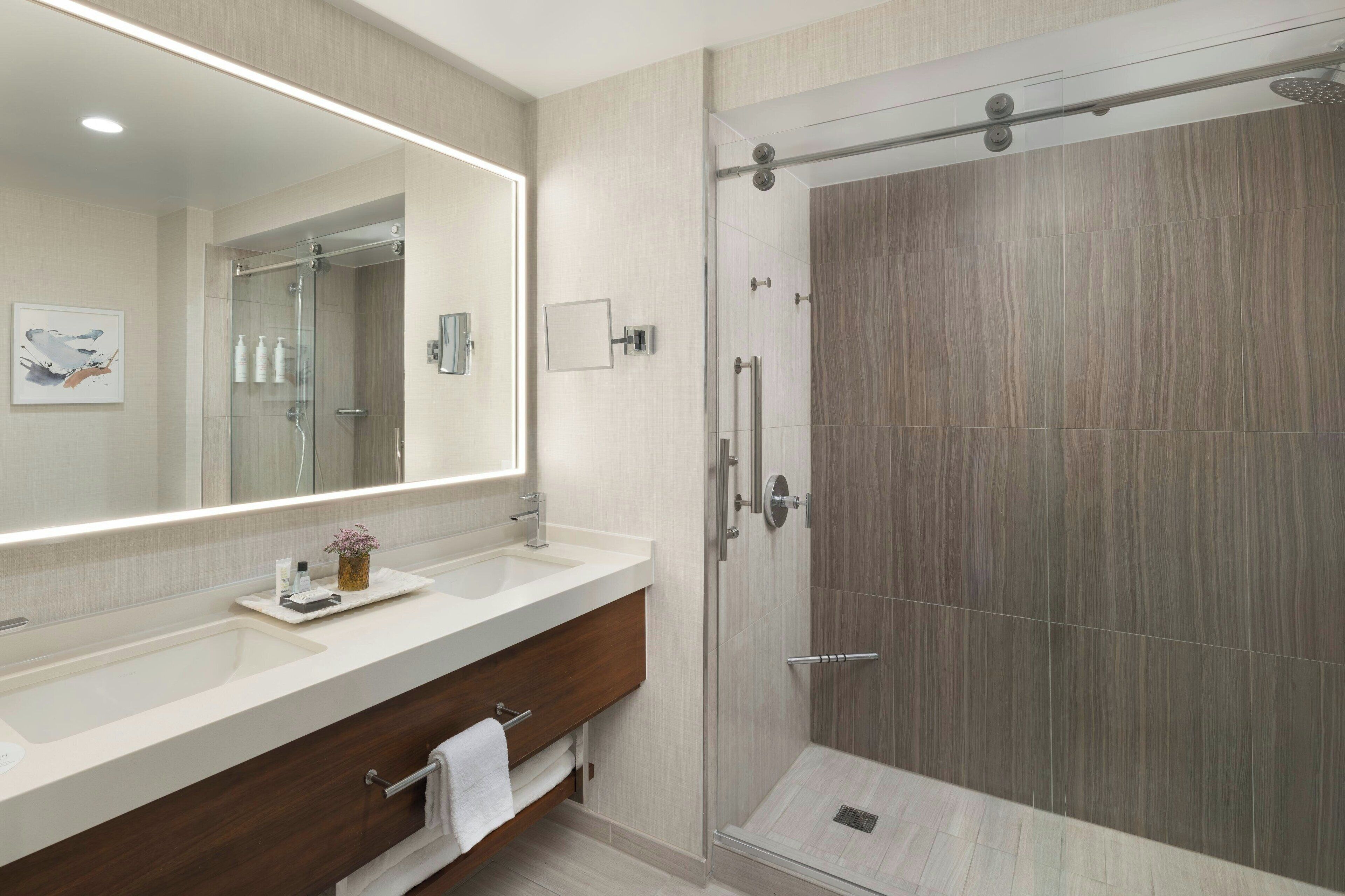 Un baño moderno con un espejo grande, dos lavamanos, una ducha y un mueble de tocador de madera.