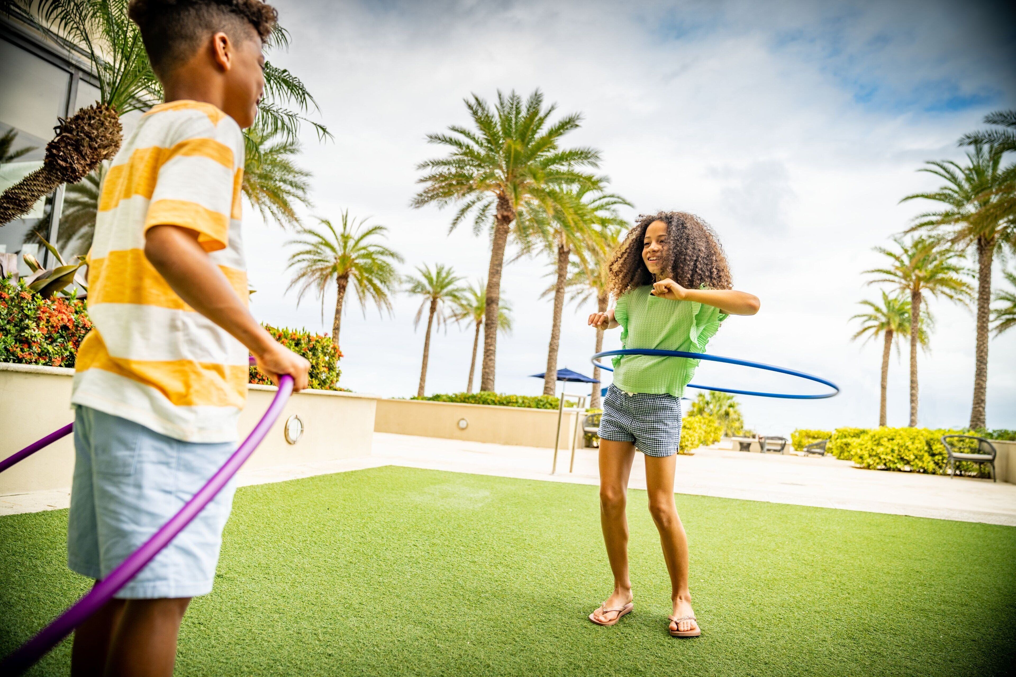 Two people playing with hula hoops on a green artificial turf area with palm trees in the background.