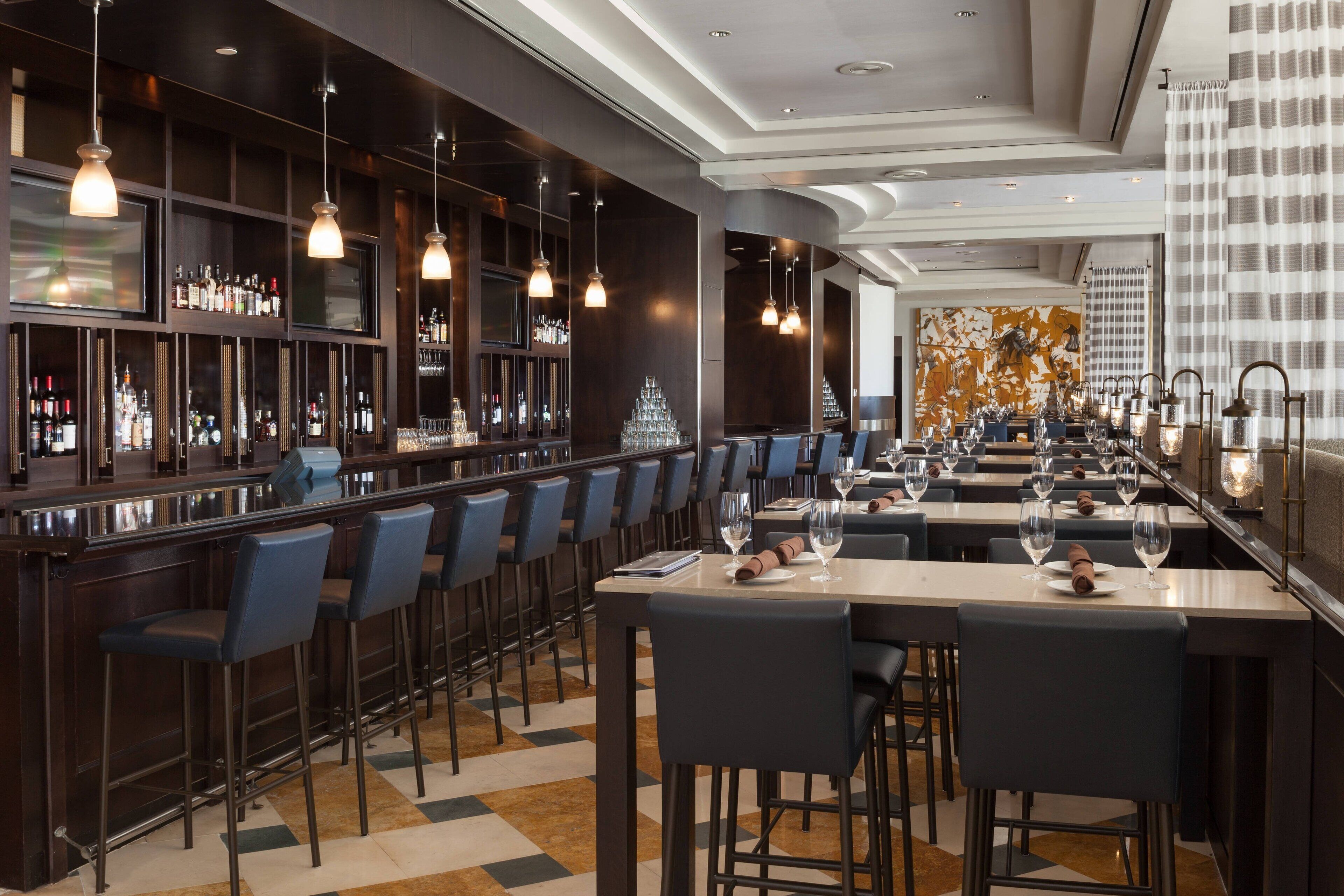 A modern bar area with a long counter, multiple bar stools, and a variety of bottles and glasses.