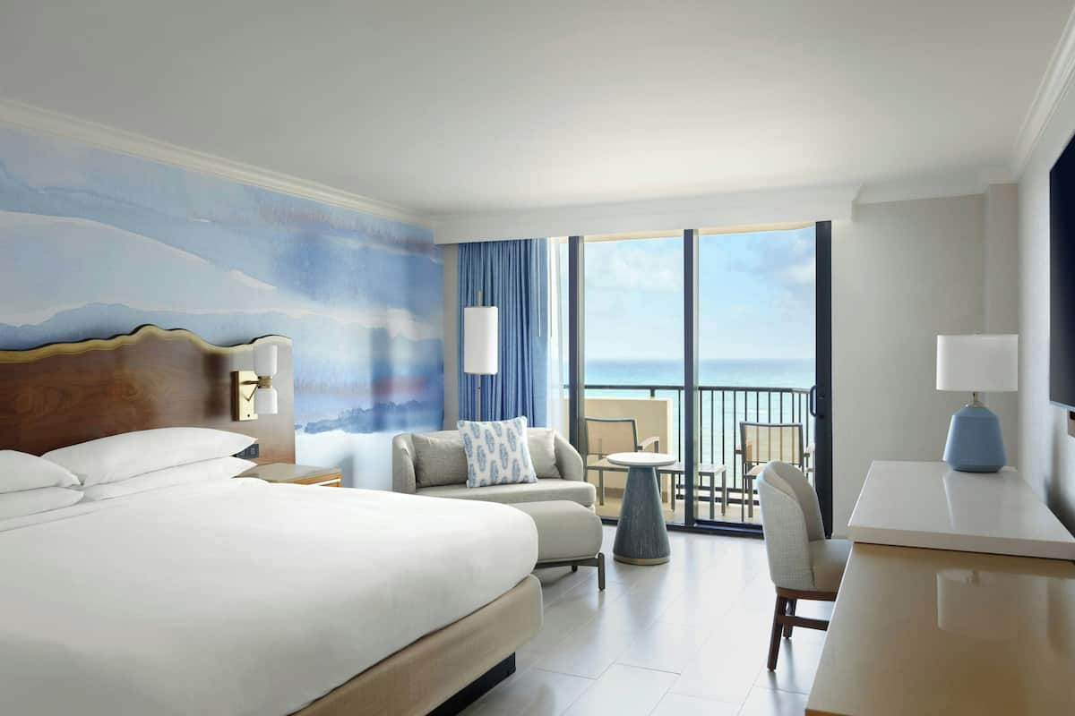 Een hotelkamer met een groot bed, een bank, een kleine tafel en een stoel. Er is een balkon met uitzicht op de oceaan.