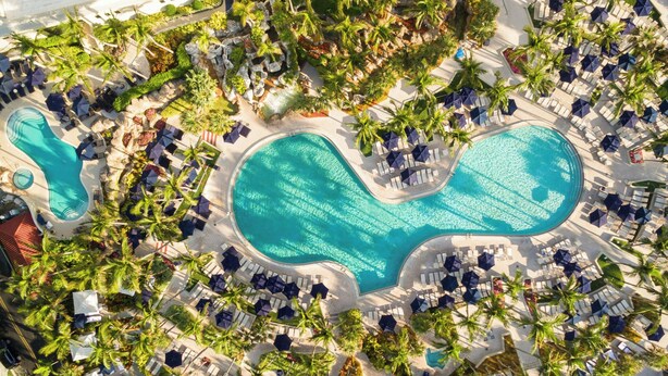 An aerial view of a resort with a large central pool surrounded by lounge areas and palm trees.