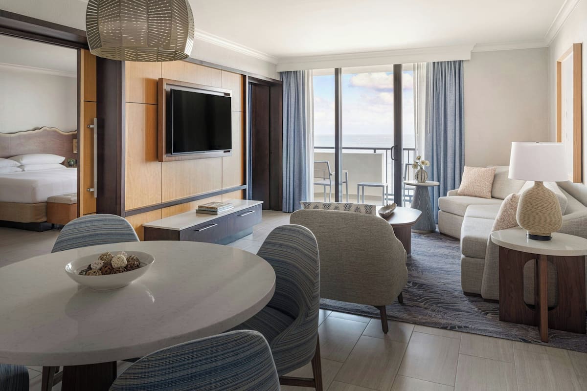 Een hotelkamer met een bed, een televisie bevestigd aan een houten paneel, een eettafel met stoelen, een bank en een balkon met uitzicht.