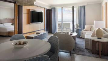 Una habitación de hotel con cama, televisión montada en un panel de madera, mesa de comedor con sillas, sofá y balcón con vistas.