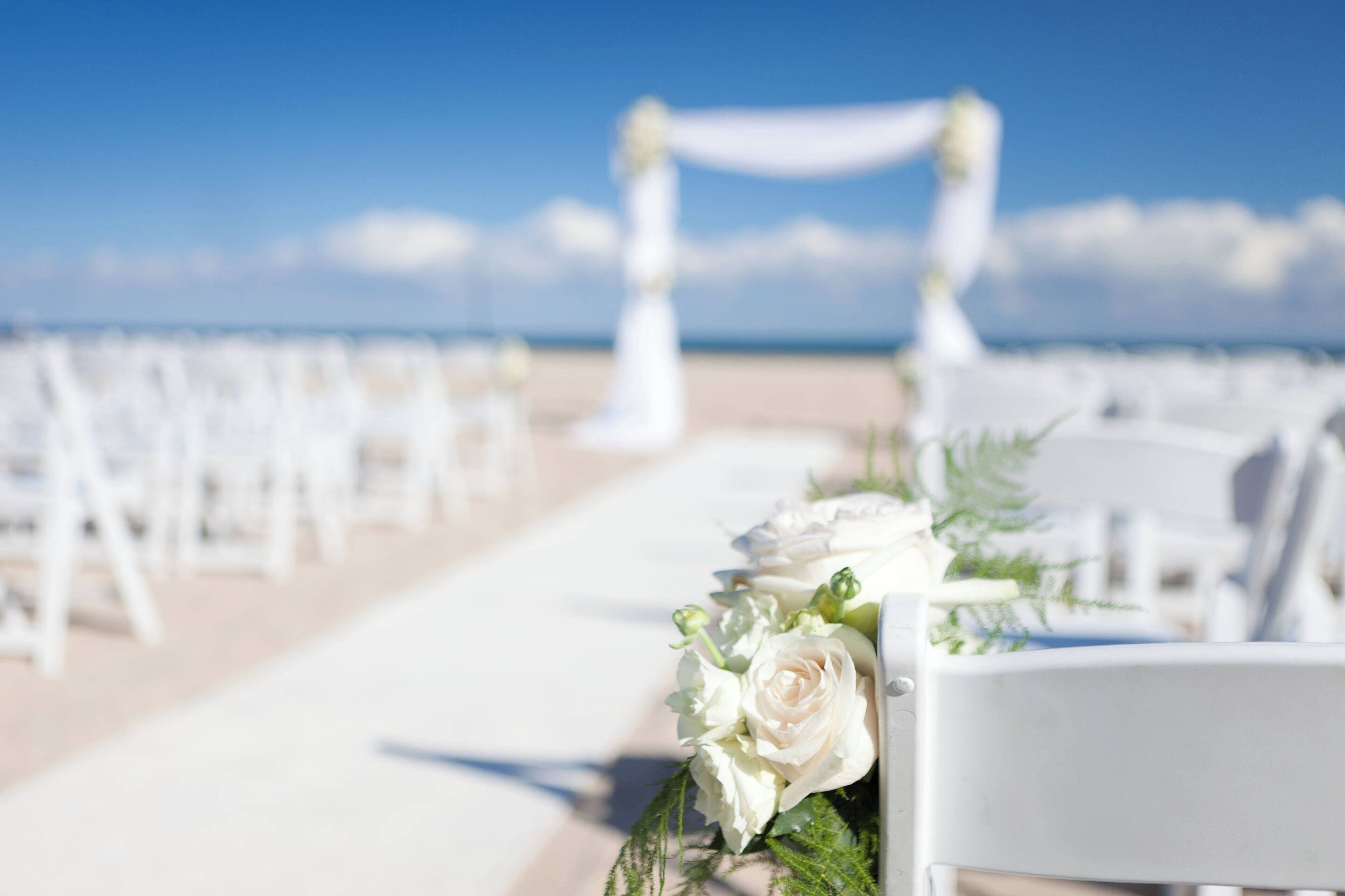 Un pasillo nupcial en la playa con sillas blancas y decoraciones florales.