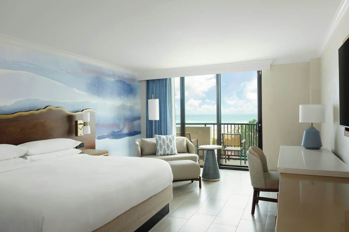Een hotelkamer met een groot bed, een bank, een tafeltje en een balkon met uitzicht op zee.