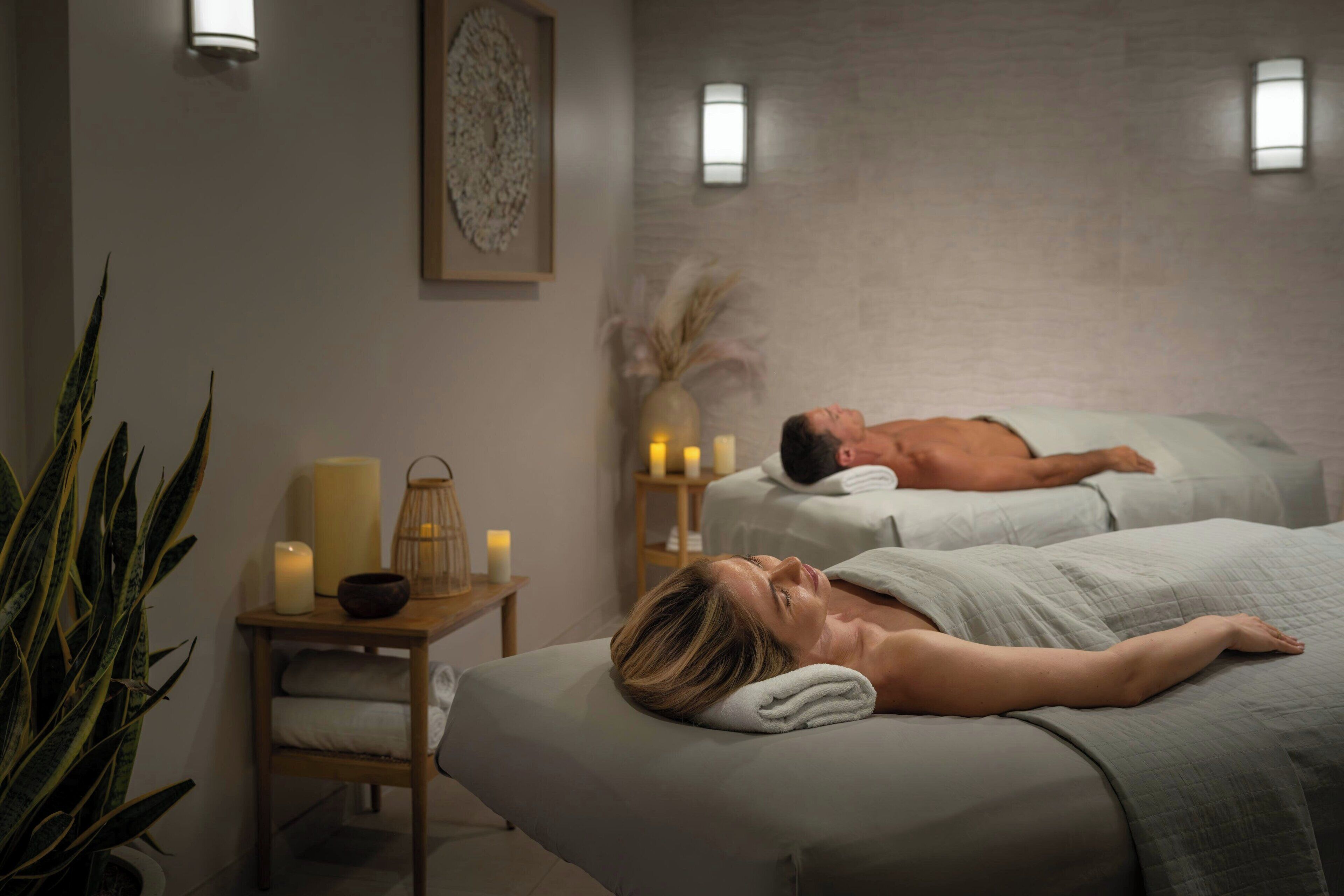 A spa room with two massage beds, a small table with candles, and a wall-mounted light fixture.