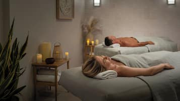 A spa room with two massage beds, a small table with candles, and a wall-mounted light fixture.