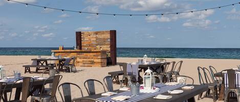An outdoor dining area with tables and chairs, a wooden structure, and a view of the sea.