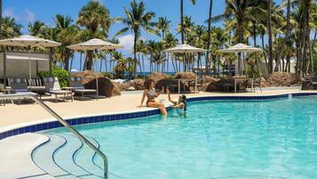 Una piscina con sillones de playa y sombrillas, rodeada de palmeras.