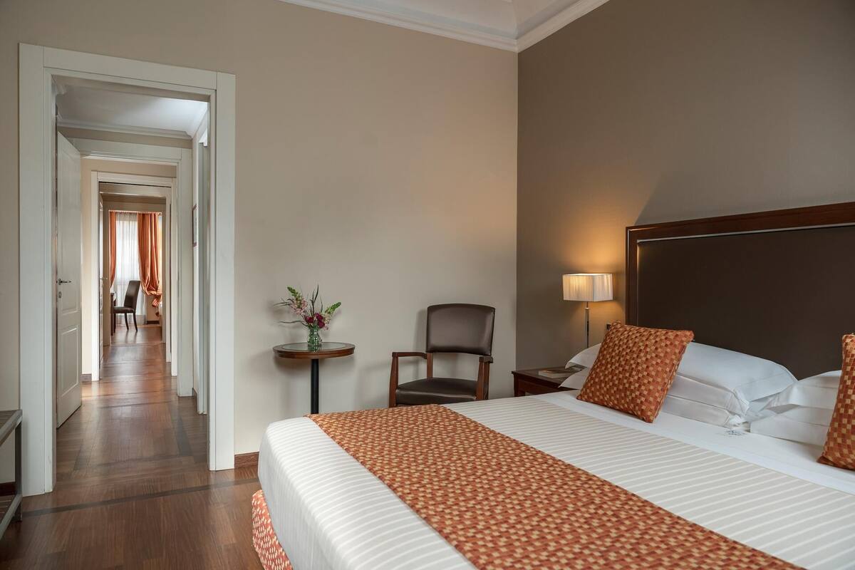 A hotel room with a large bed, a chair, a small table with a vase, and a nightstand with a lamp.