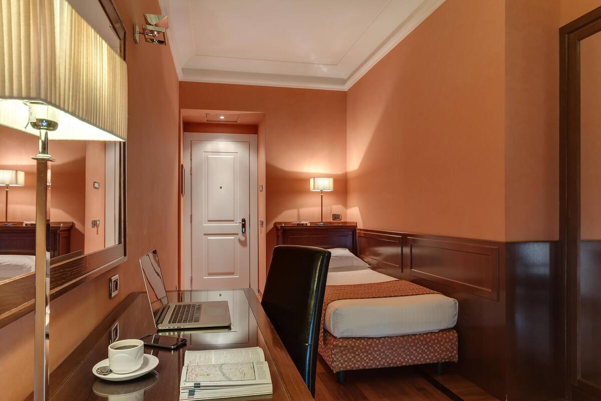 A hotel room with a bed, a desk with a laptop, a chair, a lamp, and a cup of coffee.