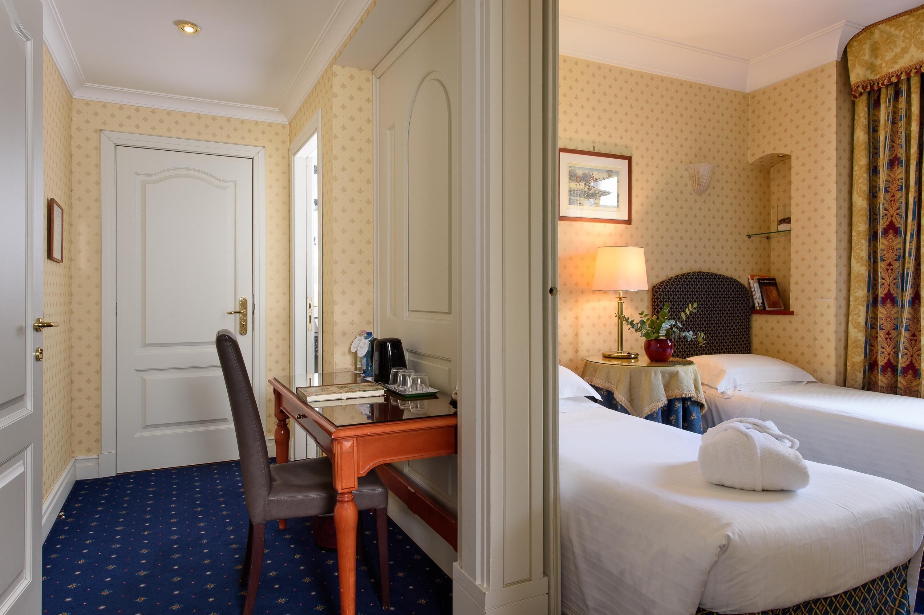 A hotel room with a desk, two beds, a chair, a lamp, and a mirror.