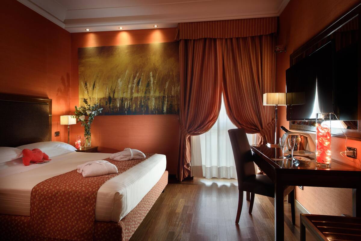 A hotel room with a large bed, a desk with a lamp, a television, and a window with curtains.