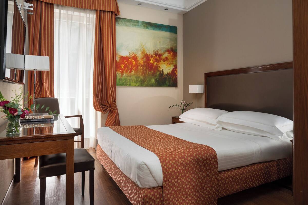 A hotel room with a bed, desk, chair, lamp, and a painting on the wall.