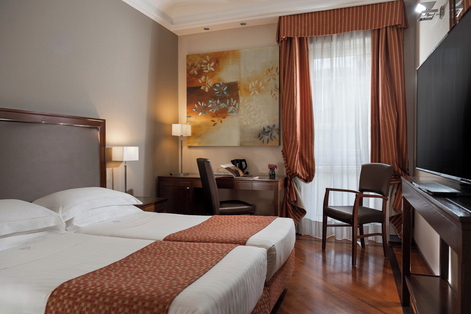 Une chambre d’hôtel avec deux lits, un bureau, une chaise, une télévision et un grand tableau au mur.