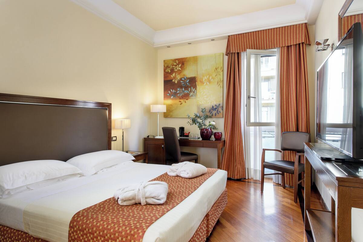 A hotel room with a large bed, a desk with a computer, a TV, and a window with curtains.