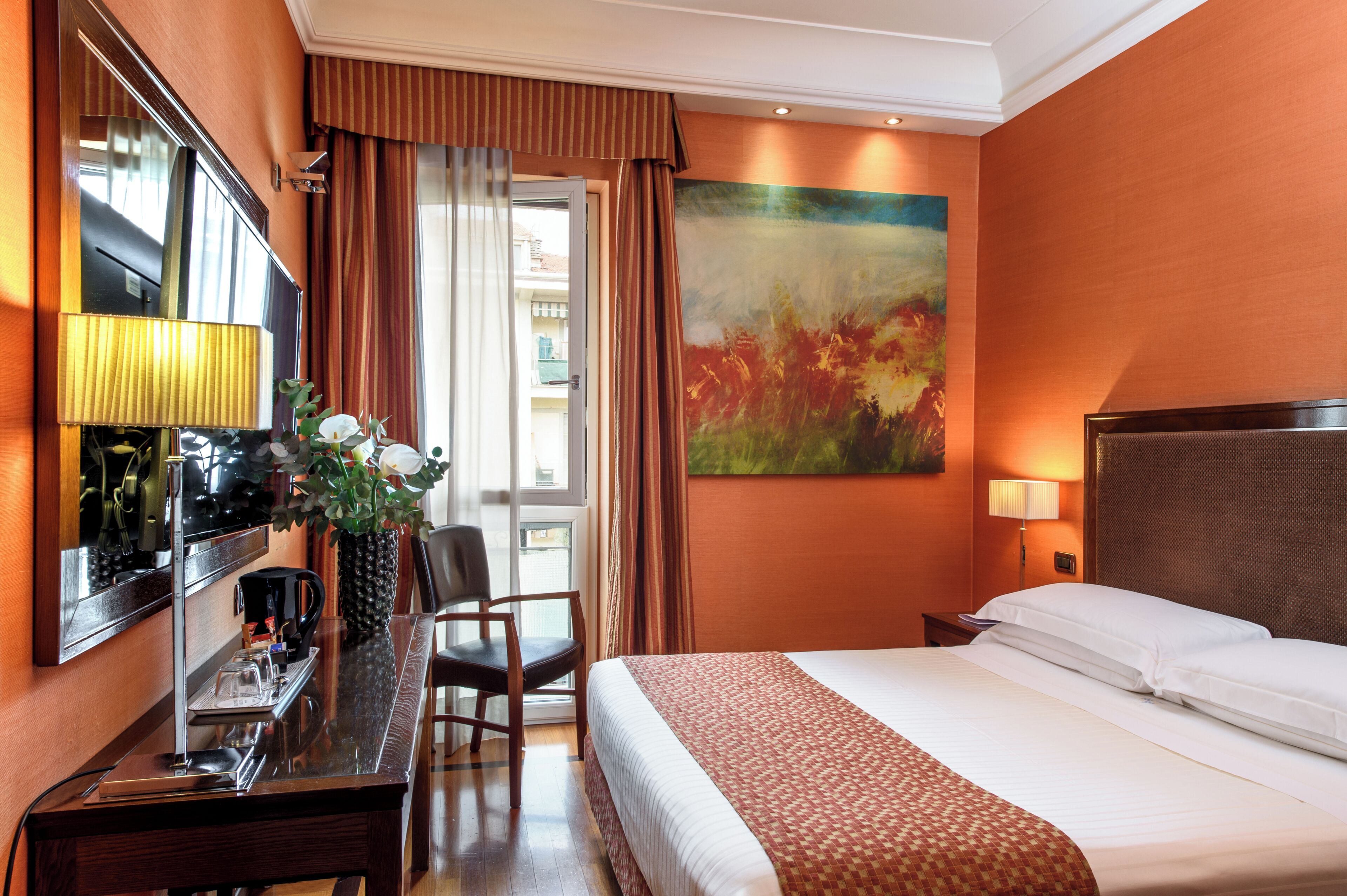 A hotel room with a bed, a desk with a lamp, a chair, a painting on the wall, and a window with curtains.