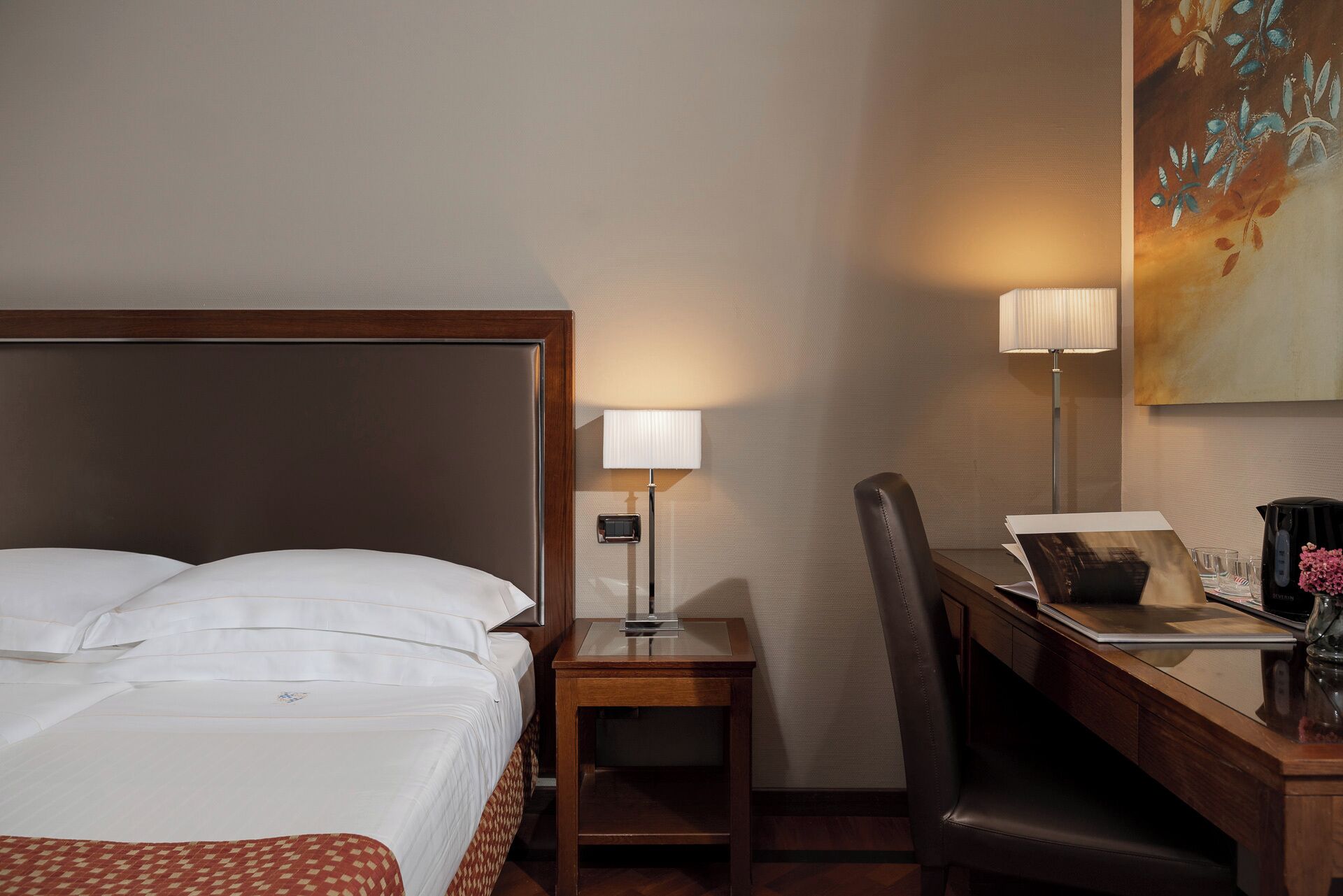A hotel room with a bed, desk, chair, lamp, and a painting on the wall.