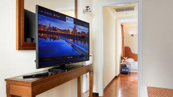 A hotel room with a flat-screen TV on a wooden stand, a bed, a nightstand, and a hallway leading to another room.