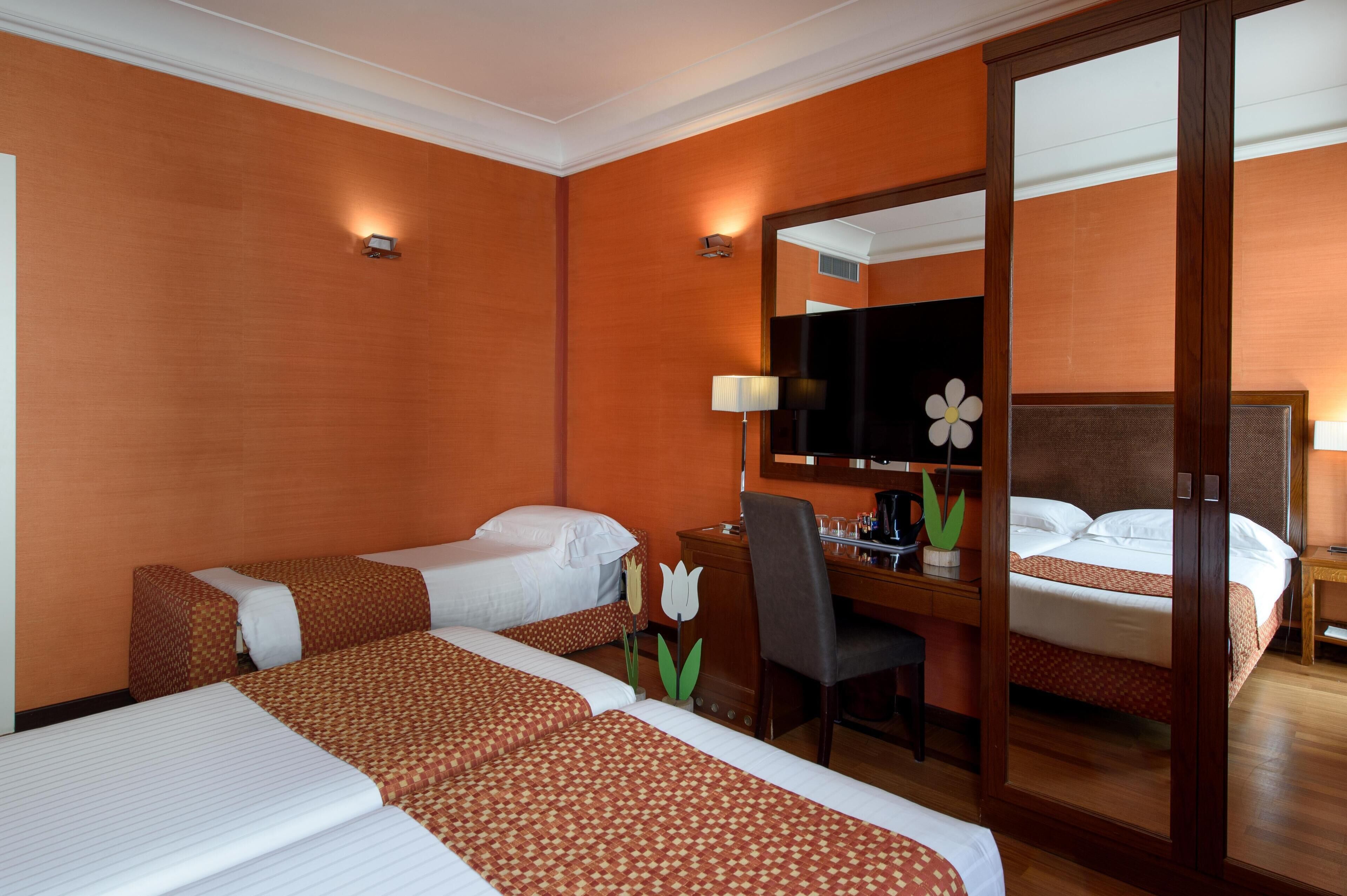 A hotel room with two beds, a desk, a chair, a mirror, and a flower decoration.
