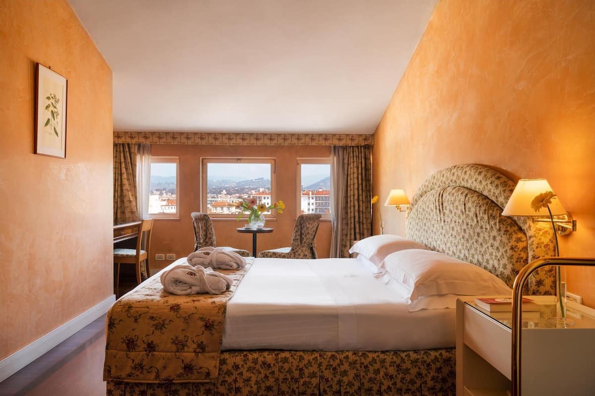 A hotel room with a bed, two chairs, a small table with a vase of flowers, and a view of the city through large windows.